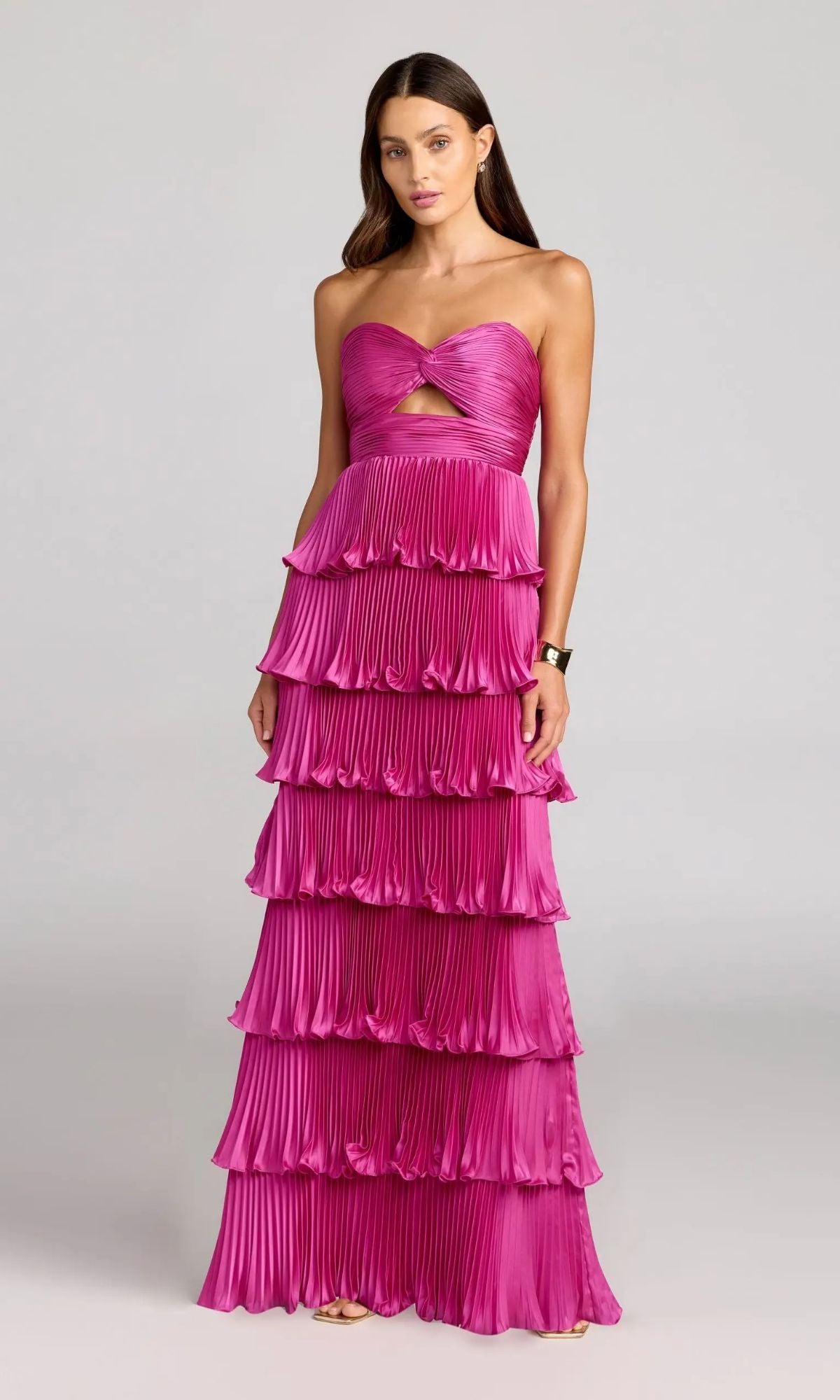 A woman poses in a fuchsia Mac Duggal 49981 long formal dress, showcasing a strapless knotted bodice cutout and tiered pleated ruffle layers against a plain gray background.